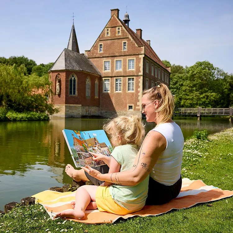 Frau und Mädchen sitzen auf der Wiese vor einem Schloss und schauen sich ein Buch an