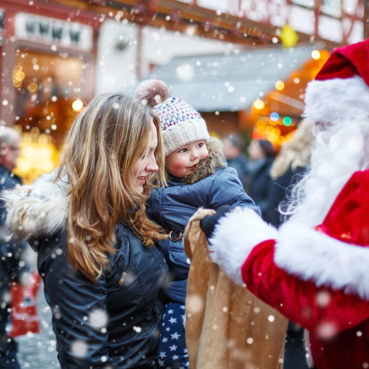 Dick eingepacktes Baby mit Pudelmütze auf dem Arm einer Frau begegnet dem Weihnachtsmann vor winterlicher Kulisse.