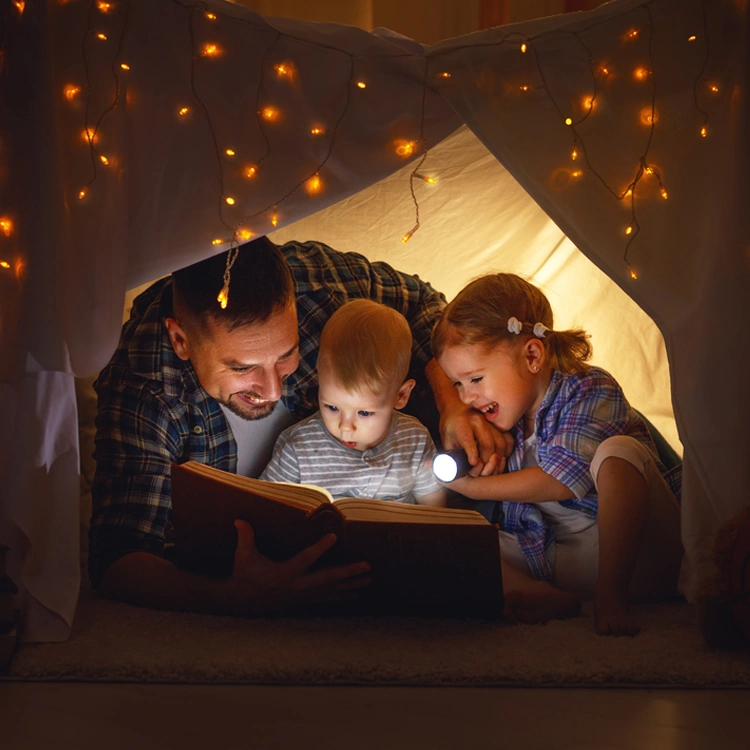 Lachende Kinder mit Vater liegen in einem beleuchteten Zelt und schauen mit einer Taschenlampe in ein Buch.