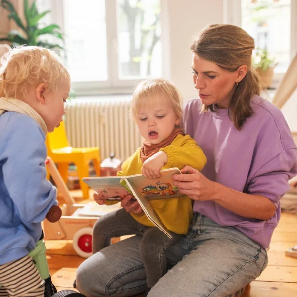 Eine Mutter liest im Kinderzimmer mit ihren Kindern ein Buch. Eines sitzt auf ihrem Schoß, das andere auf einem Dreirad und schaut interessiert in das Buch.