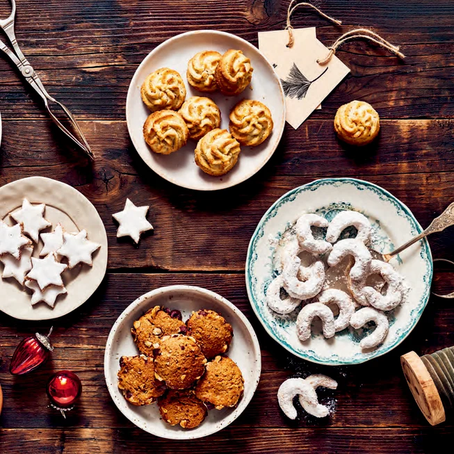 Verschiedene Kekssorten attraktiv angerichtet auf Tellern auf einem alten Tisch. Dazu weihnachtliche Dekoration.